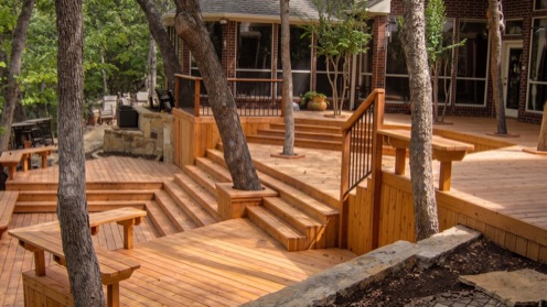 Multi-tiered wooden deck structure surrounded by trees, featuring stairs, railings, and multiple levels